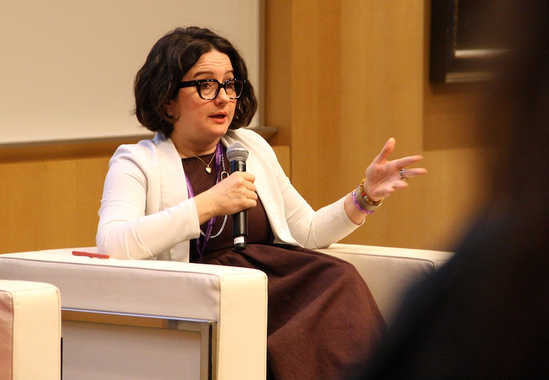 A woman in a white jacket and glasses holds a microphone.