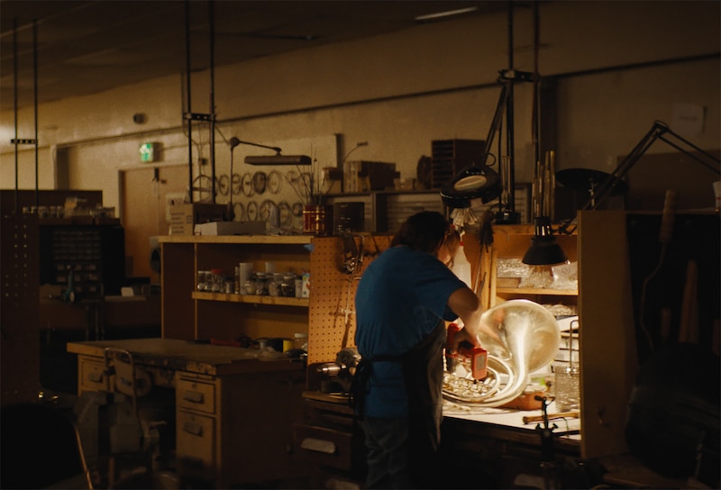 A person stands at a workbench in a darkly lit room with tools along the wall in the background.