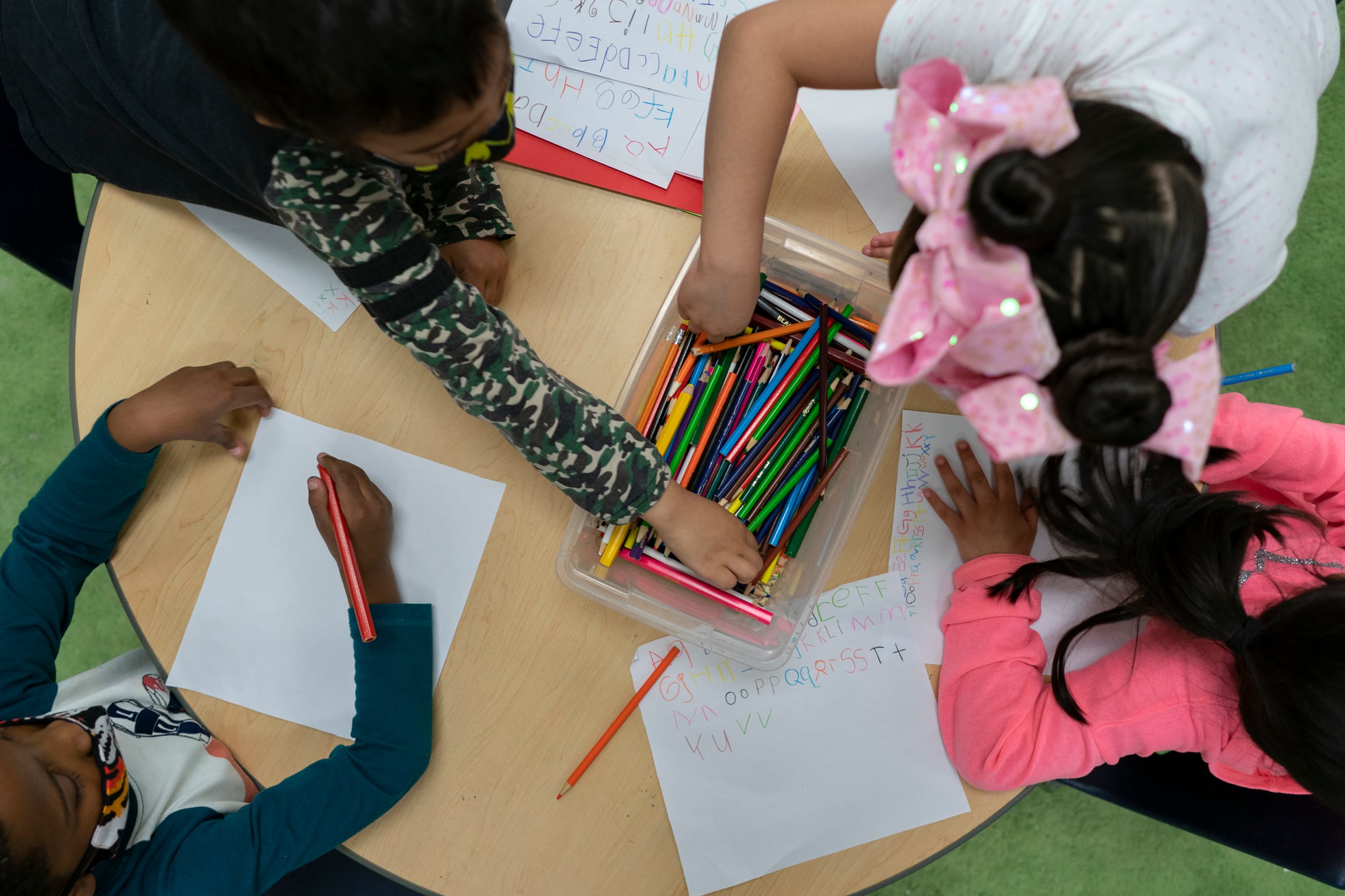 Aerial-style view of the top of four young children’s heads as they practice writing the letters of the alphabet using colored pencils from a shared container in the middle of the four.