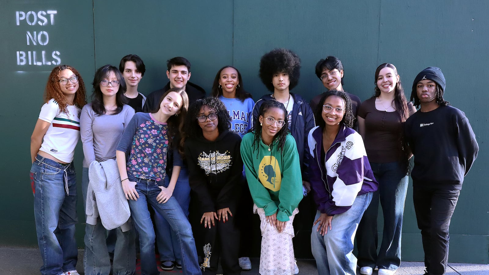 A group of high school students pose for a portrait in front of a green wall outside.