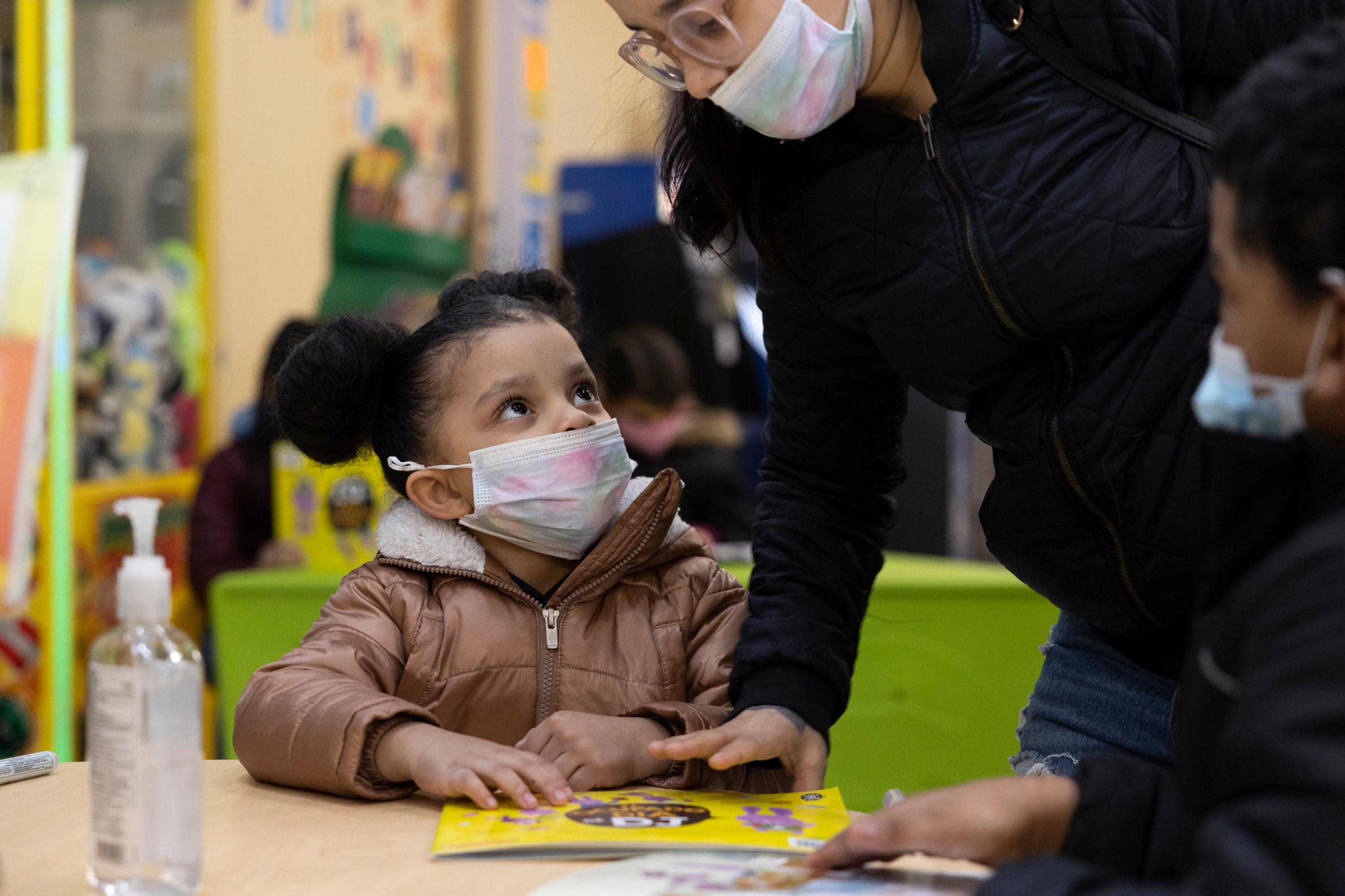 A woman in a black jacket helps a young girl as she’s reading a children’s book.