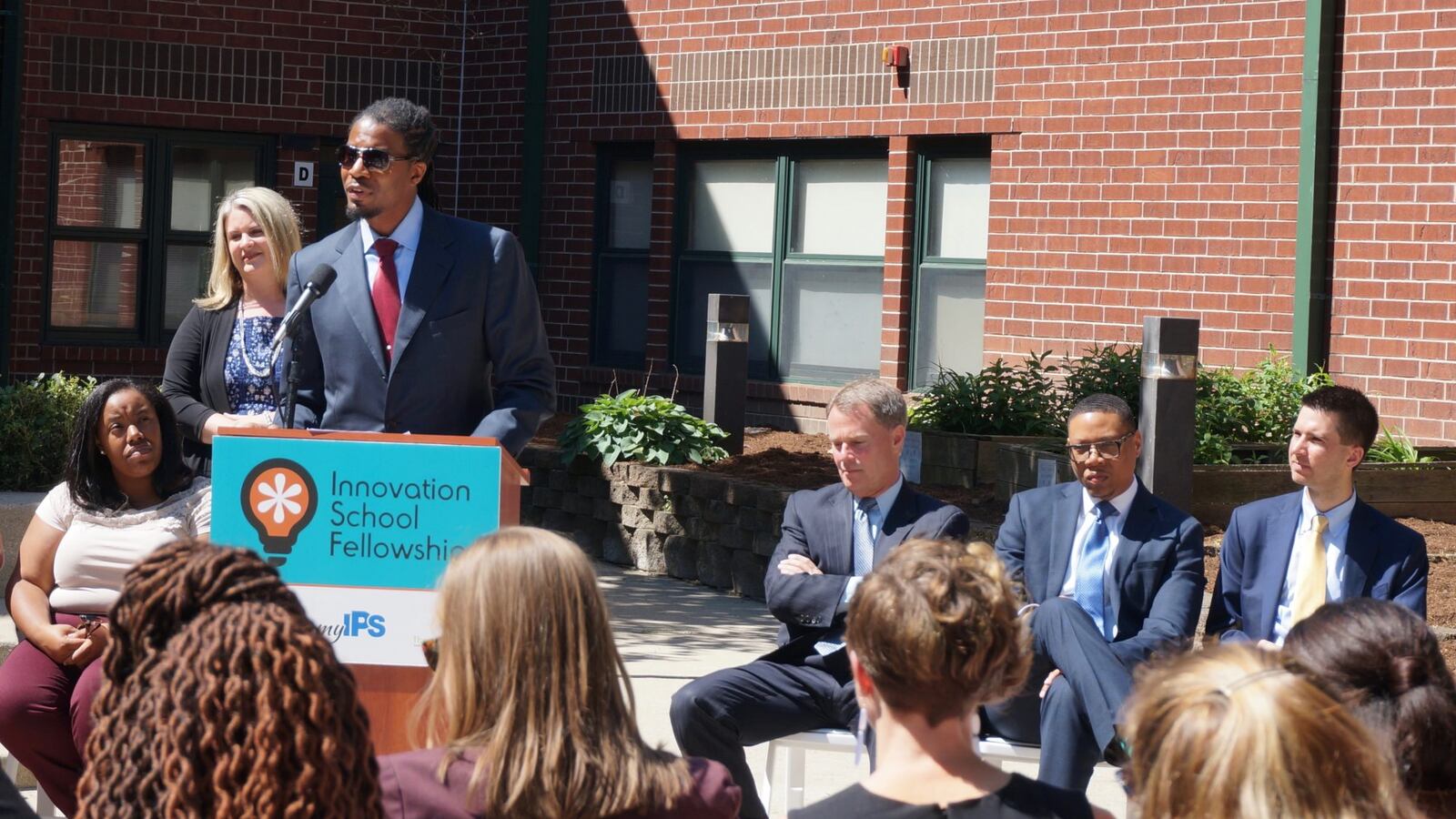 A person wearing a suit speaks from a podium while others sit in chairs beside him and in the crowd. They are outside in the sun and a red brick building in the background.