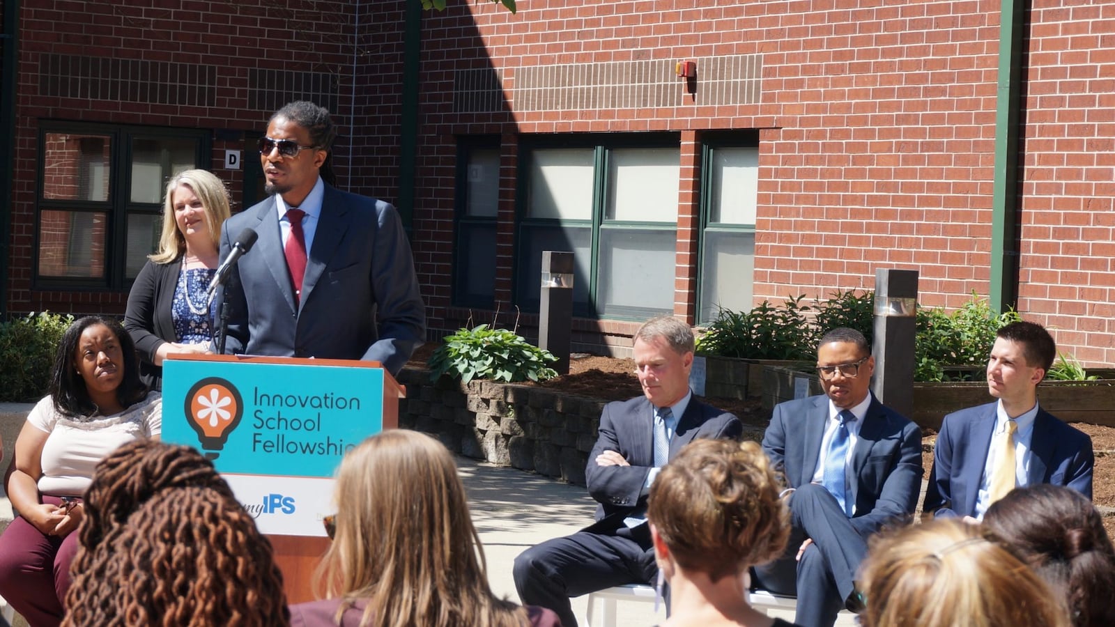 A person wearing a suit speaks from a podium while others sit in chairs beside him and in the crowd. They are outside in the sun and a red brick building in the background.