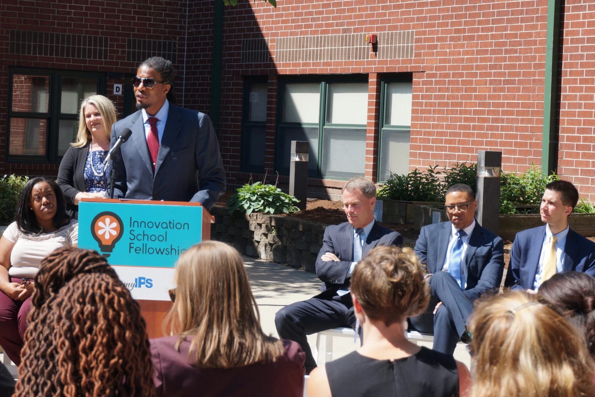 A person wearing a suit speaks from a podium while others sit in chairs beside him and in the crowd. They are outside in the sun and a red brick building in the background.