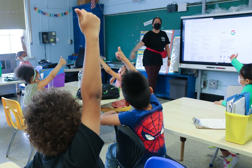 Children sitting at tables give a thumps up to a teacher in the background