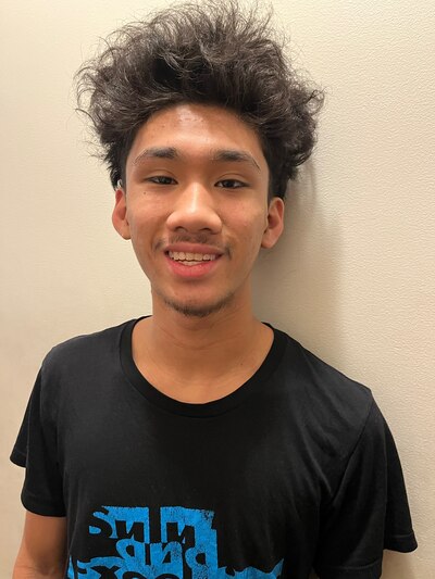 A photograph of a high school senior student with dark hair and wearing a black t-shirt poses for a portraits against a white wall.
