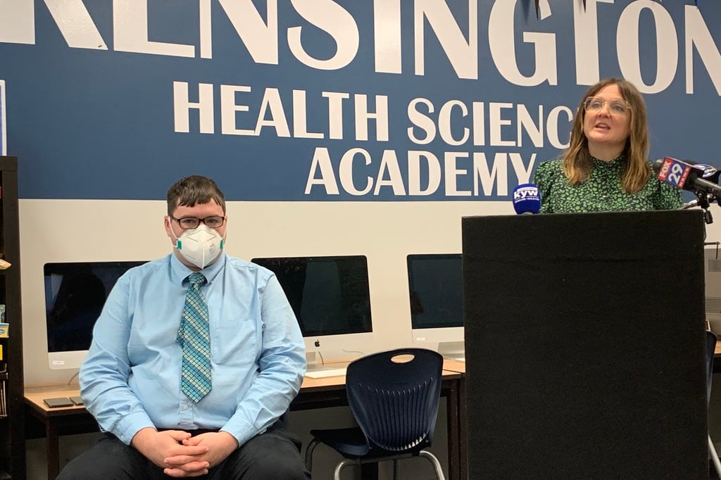A man wearing glasses, a face mask, a blue shirt, dark pants, and a tie sits as a woman in a green and black shirt speaks into microphones at a podium. A blue school sign hangs behind them.