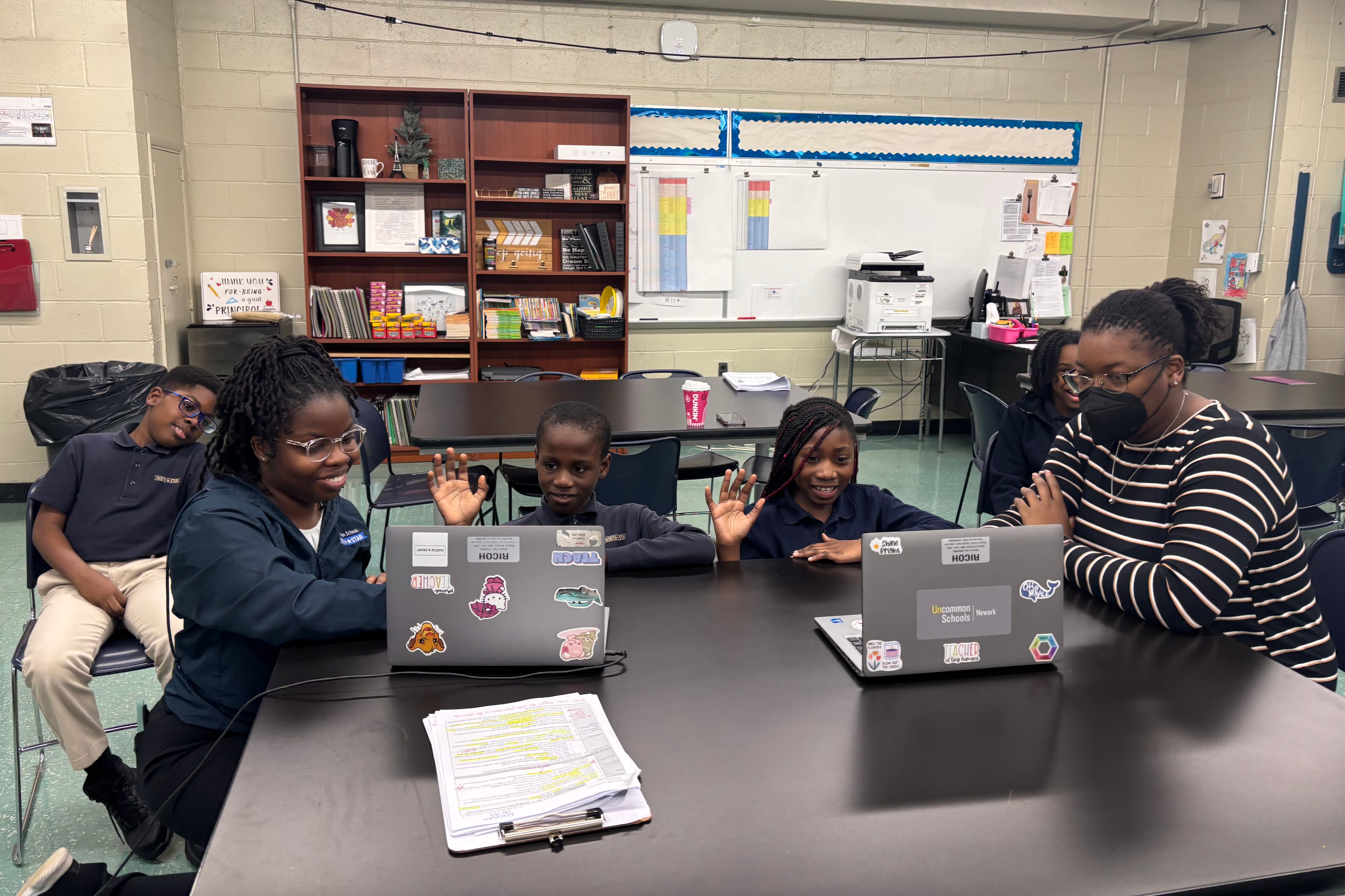 A woman with glasses kneels on the left side of a table where two students are looking at laptops.