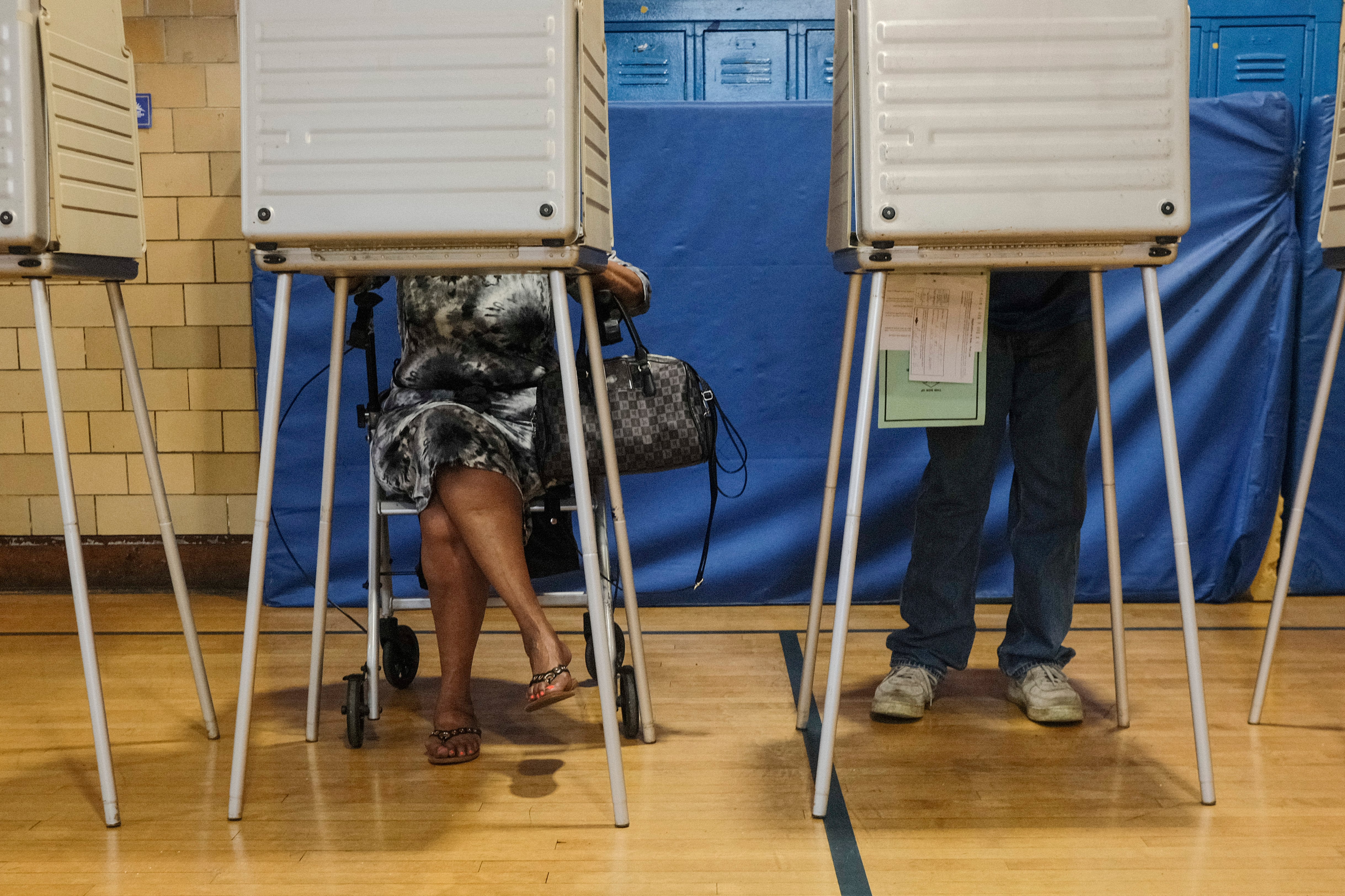 Two people cast their ballots in voting booths. Their top halves are behind a voting screen and their legs are visible beneath the table. The voter on the left is sitting in a chair with legs crossed. The voter on the right is standing.