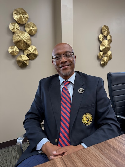 A photograph of a Black man in a dark suit and red tie posing for a photograph while sitting at a desk.