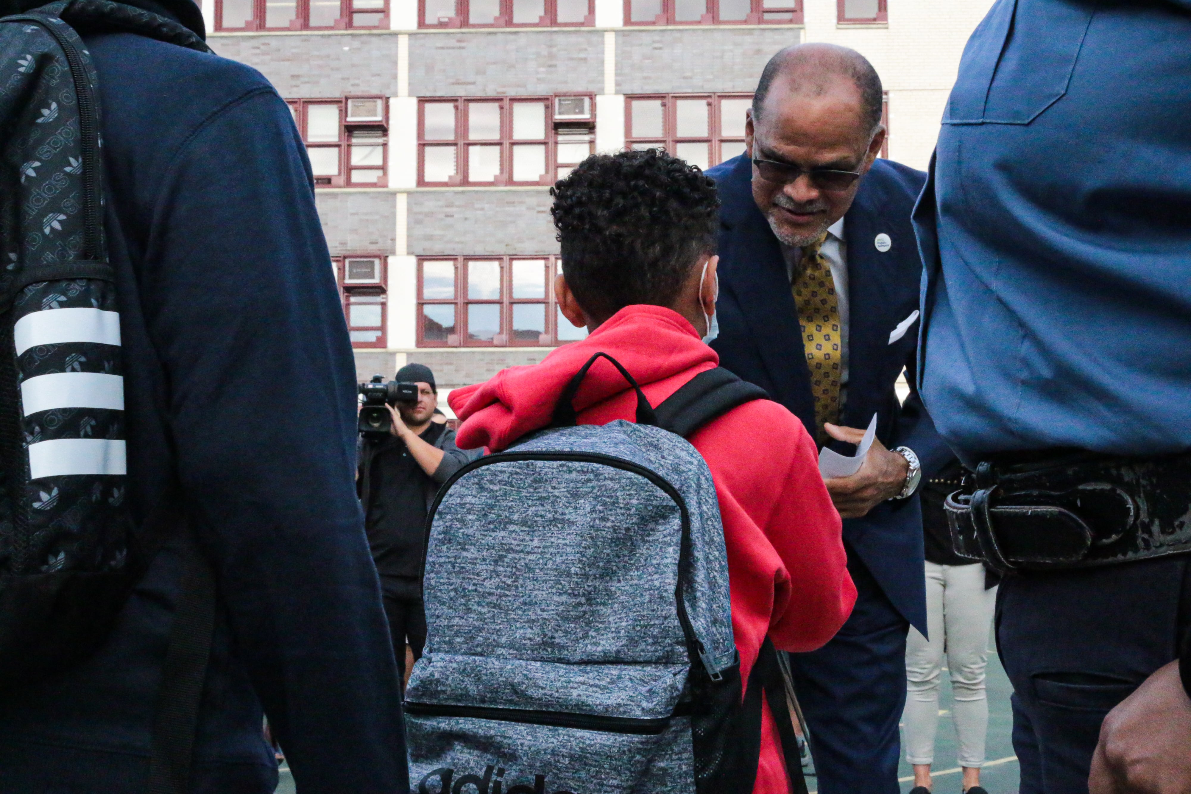 A man in a suit bends down to talk to a student wearing a backpack. The student is facing away.