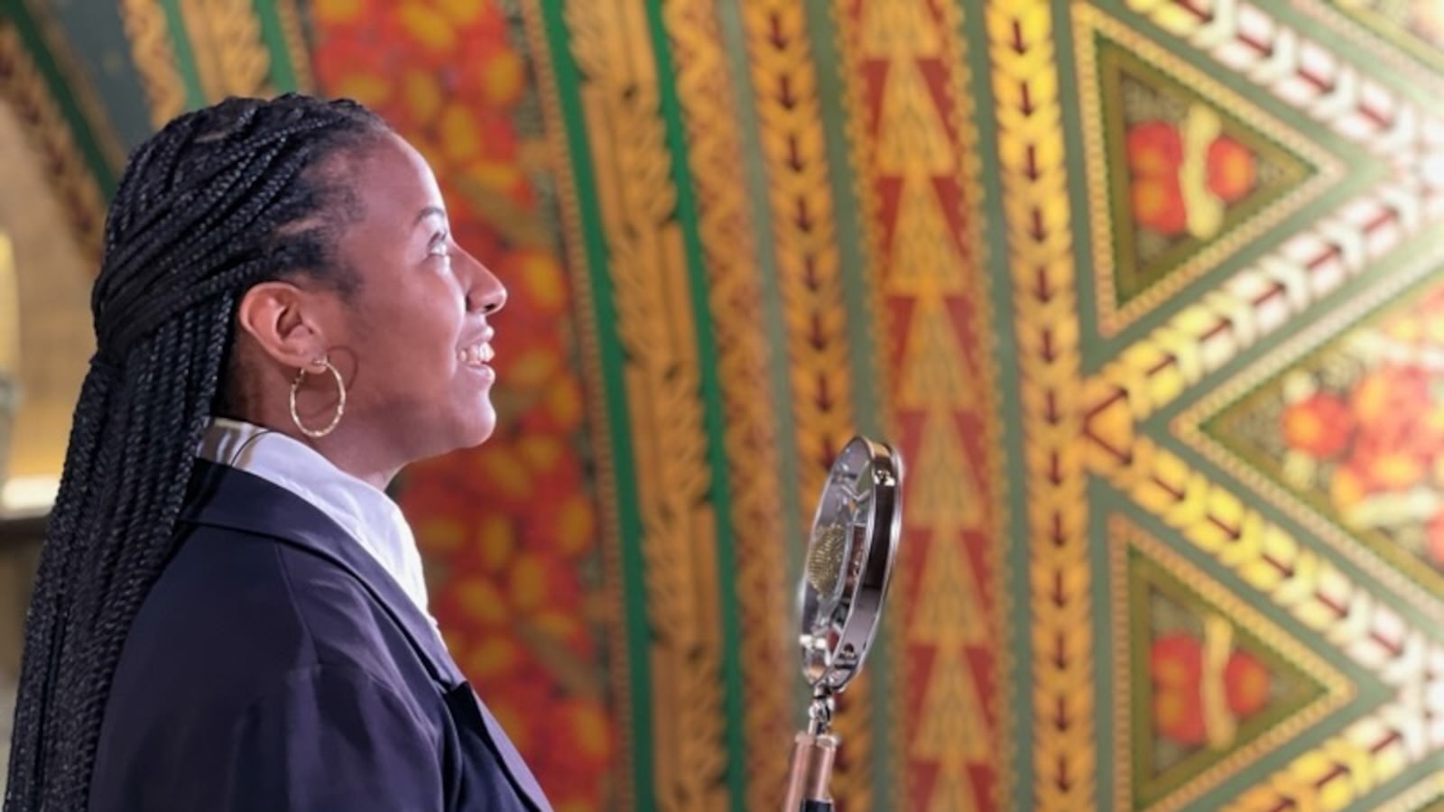 The profile of a young woman speaking in front of a microphone. She wears a white shirt and a black blazer.