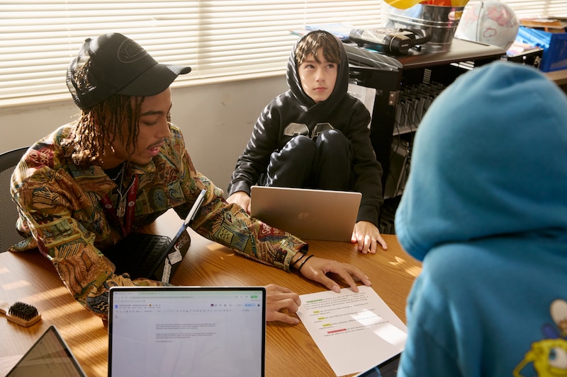A male student in a black cap works with students at a table.