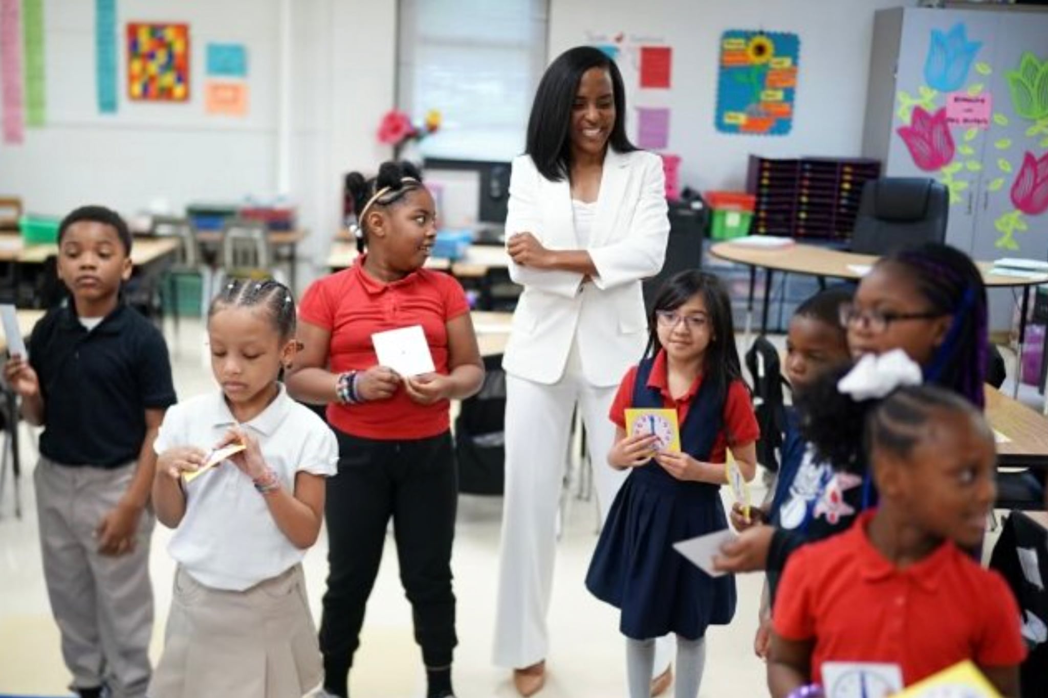 An adult woman wearing an all white suit stands in a classroom next to a group of young elementary students.