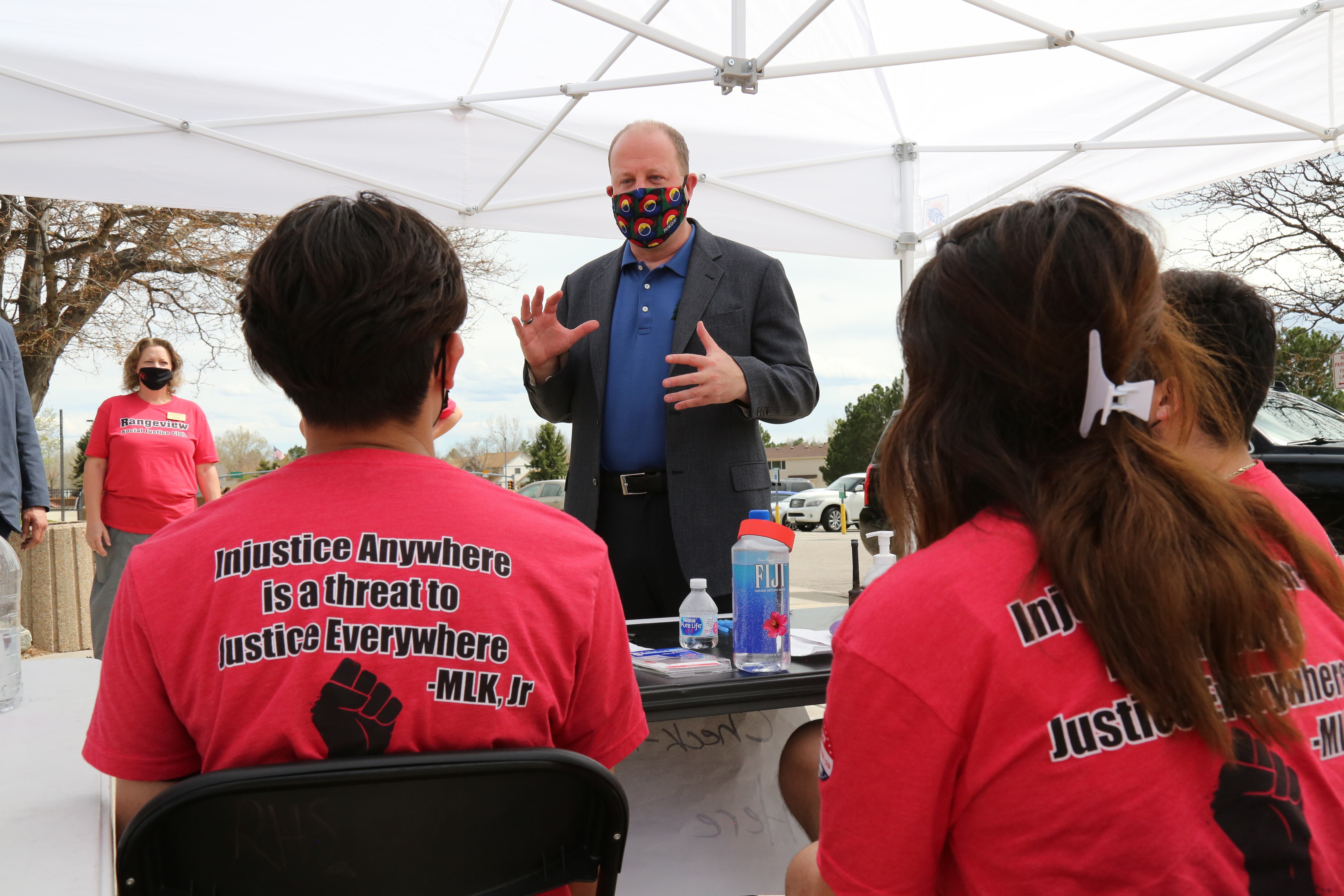 Gov. Jared Polis, wearing a mask, stands in the center of the image. He’s talked to two seated students in red t-shirts, their backs to the camera.