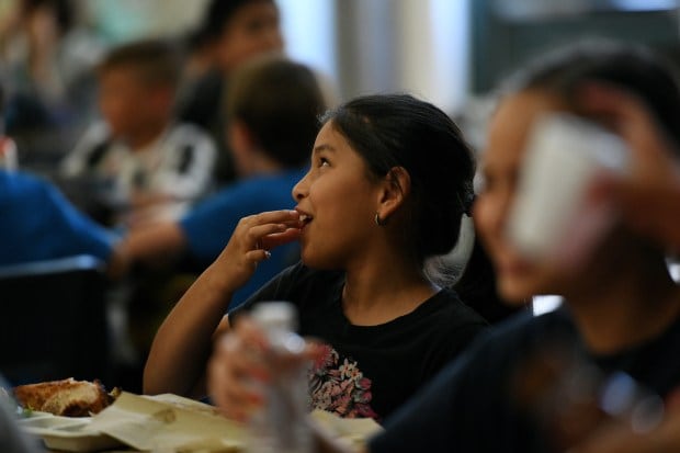 A smiling girl with dark hair pops some food in her mouth as she sits with friends in a crowded lunchroom.