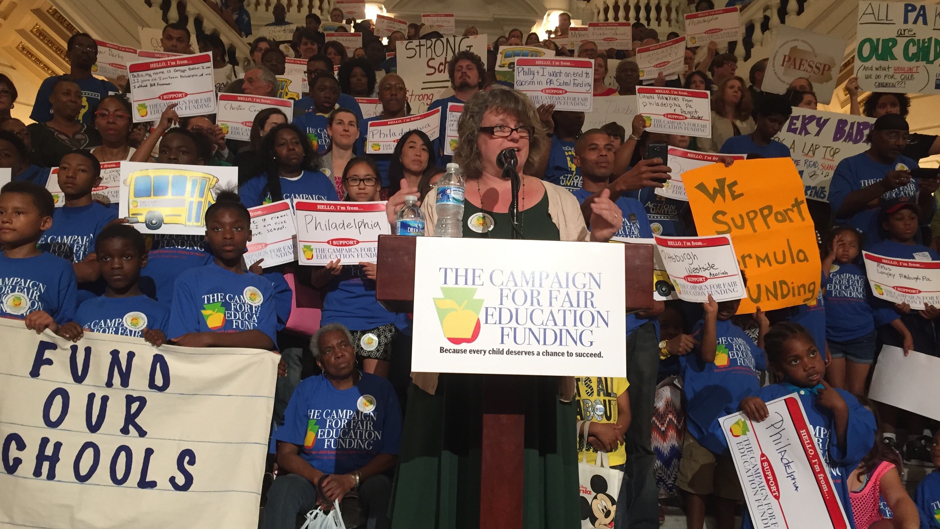 A woman in a green dress stands at a podium and speaks into a microphone while several people hold up signs behind her. The podium has a placard that says “The Campaign For Fair Education Funding.” One sign to the left says “Fund Our Schools.”