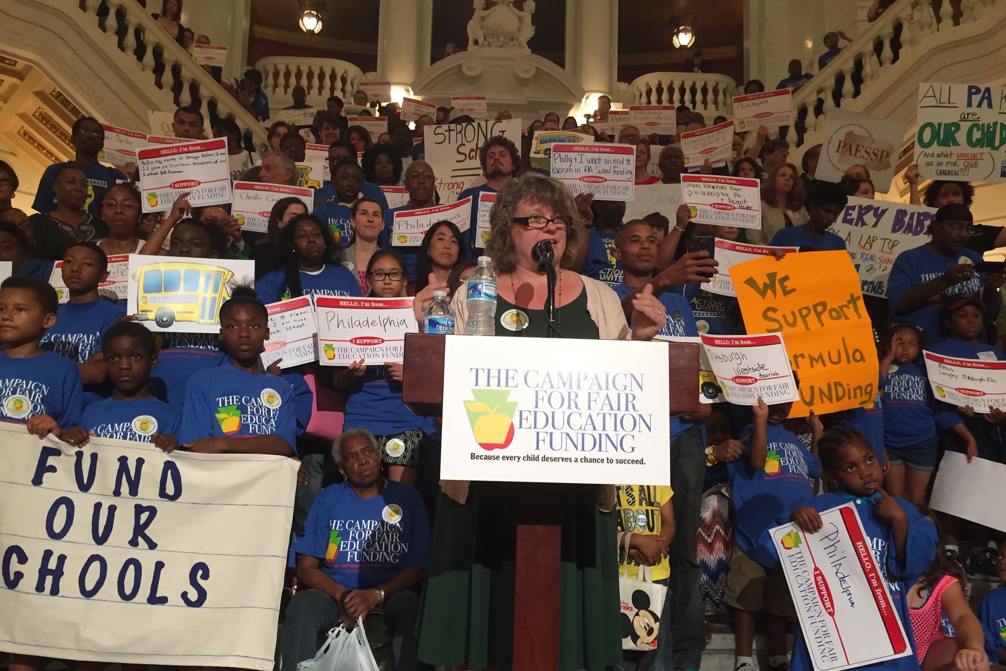 A woman in a green dress stands at a podium and speaks into a microphone while several people hold up signs behind her. The podium has a placard that says “The Campaign For Fair Education Funding.” One sign to the left says “Fund Our Schools.”