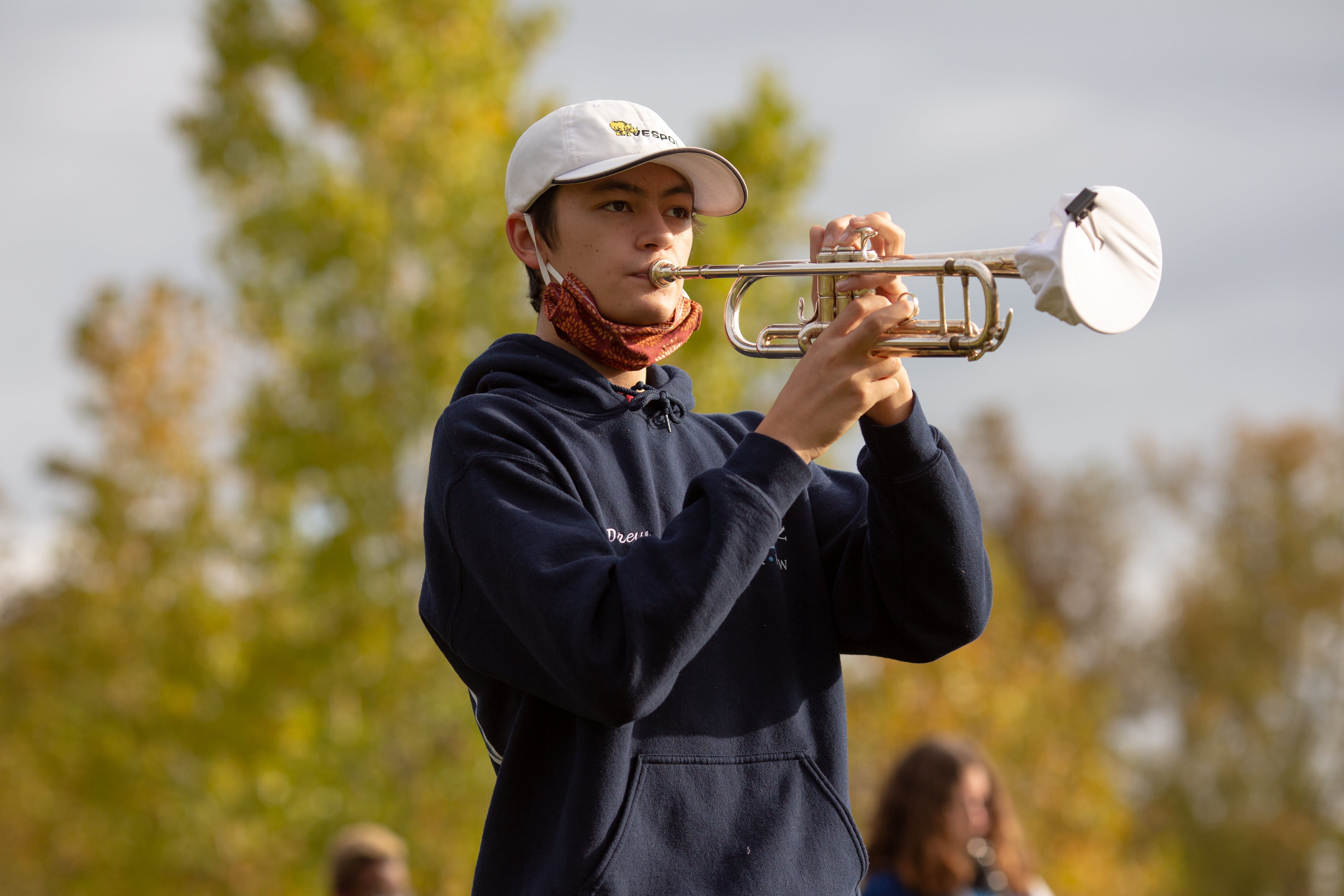 A student with a red mask below his chin plays a trumpet outside. Trees are in the background.