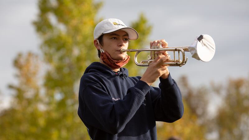 A student with a red mask below his chin plays a trumpet outside. Trees are in the background.