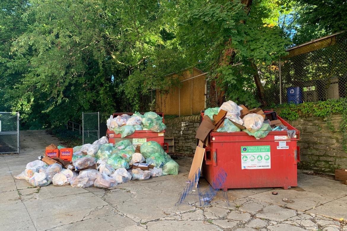 Dozens of trash bags sit next to a red dumpster filled with more trash bags.