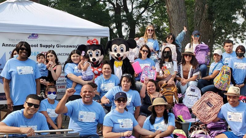 Denver School Board candidate Jose Silva, center left, poses with about two dozen people during a backpack and school supply drive. Many are wearing sky blue T-shirts and two are dressed as Mickey Mouse.