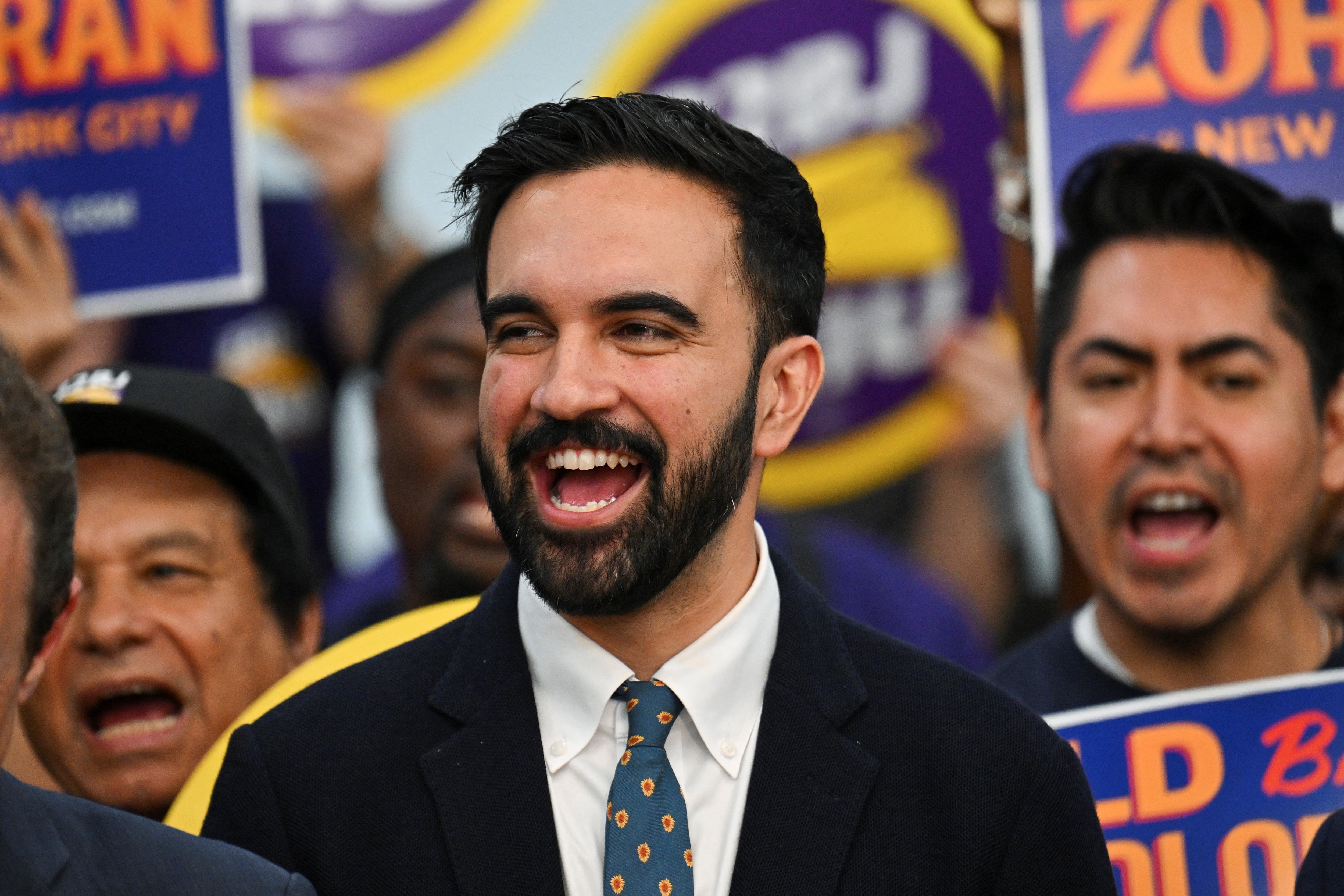 A man in a dark suit jacket smiles surrounded by other people holding signs.