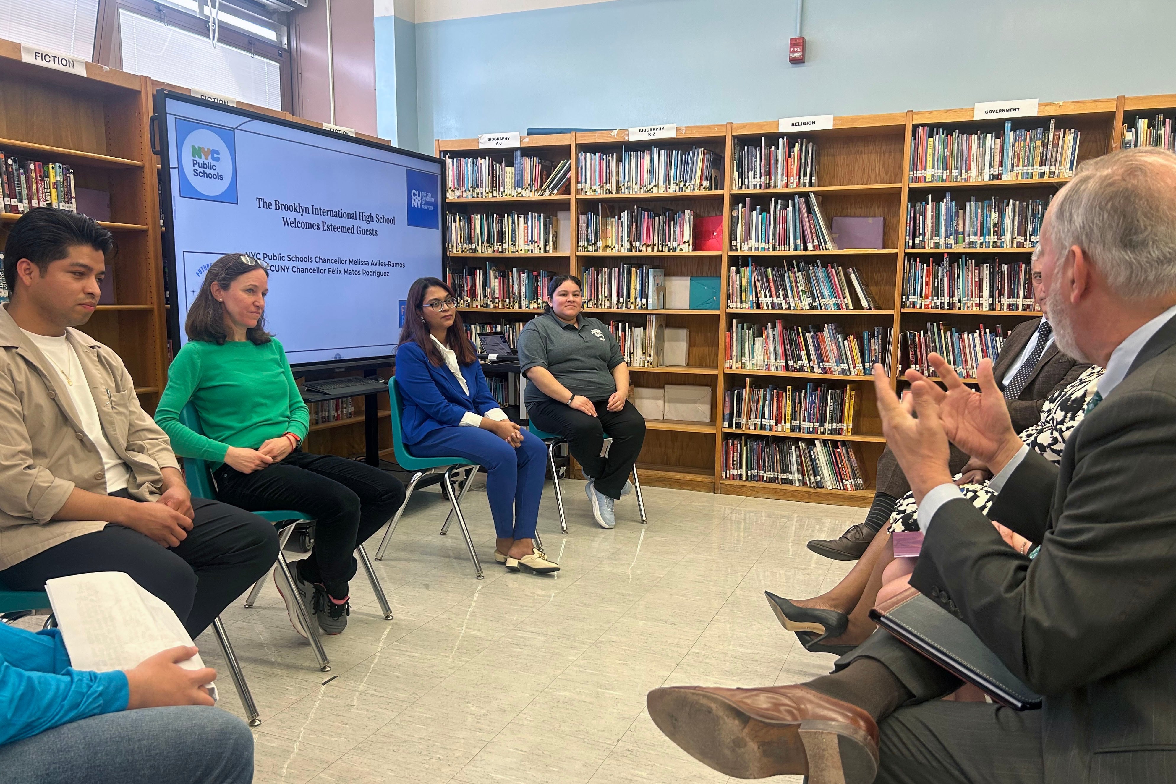 A group of adults sit in chairs in a school library facing each other during a discussion.