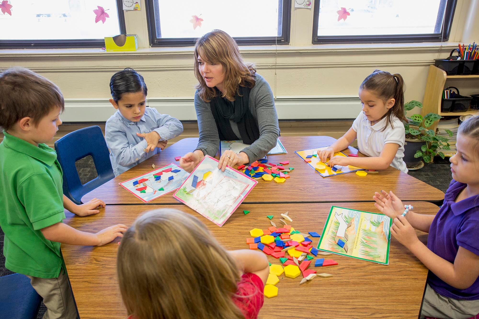 A woman with light brown hair sits at a round table teaching five students in a classroom with large windows behind them.