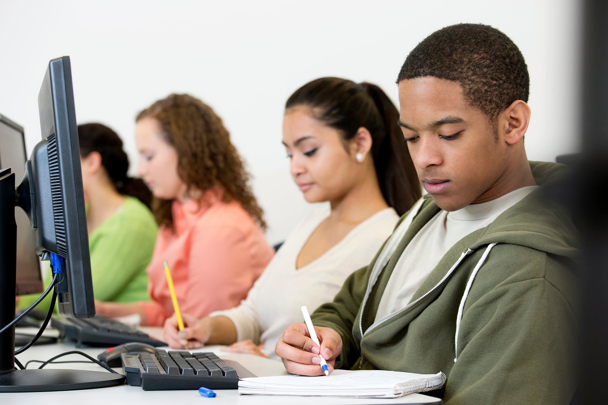 Five students sit in a row with computers in front of them and a pen and paper on the desk. The background is a plain white wall.