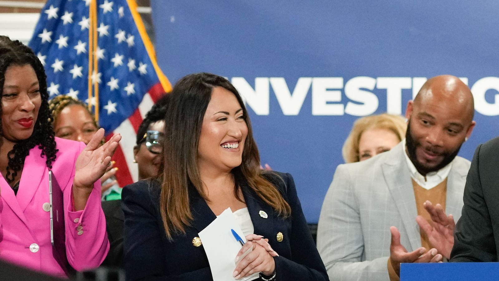 two women wearing suits stand in front of a group of people.