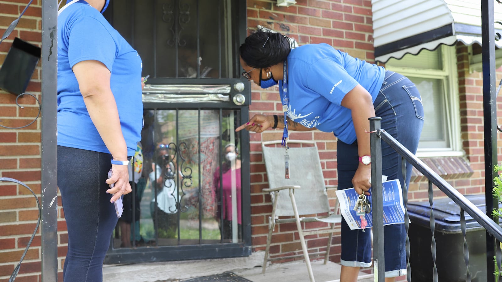 Two women wearing blue T-shirts and jeans stand on the front porch of a home and speak with a person through a glass door.