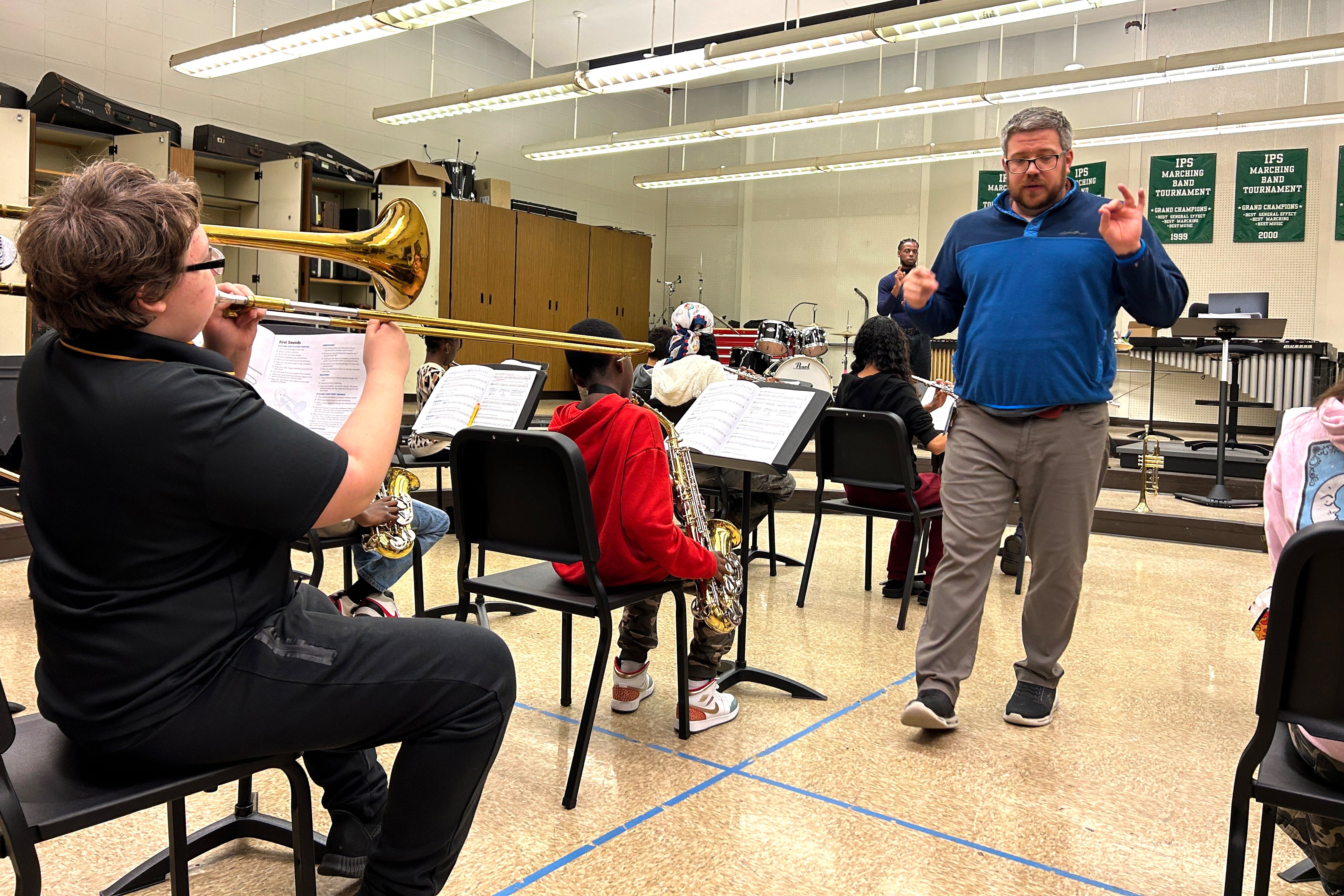 A man in a blue shirt leads a group of students playing music.