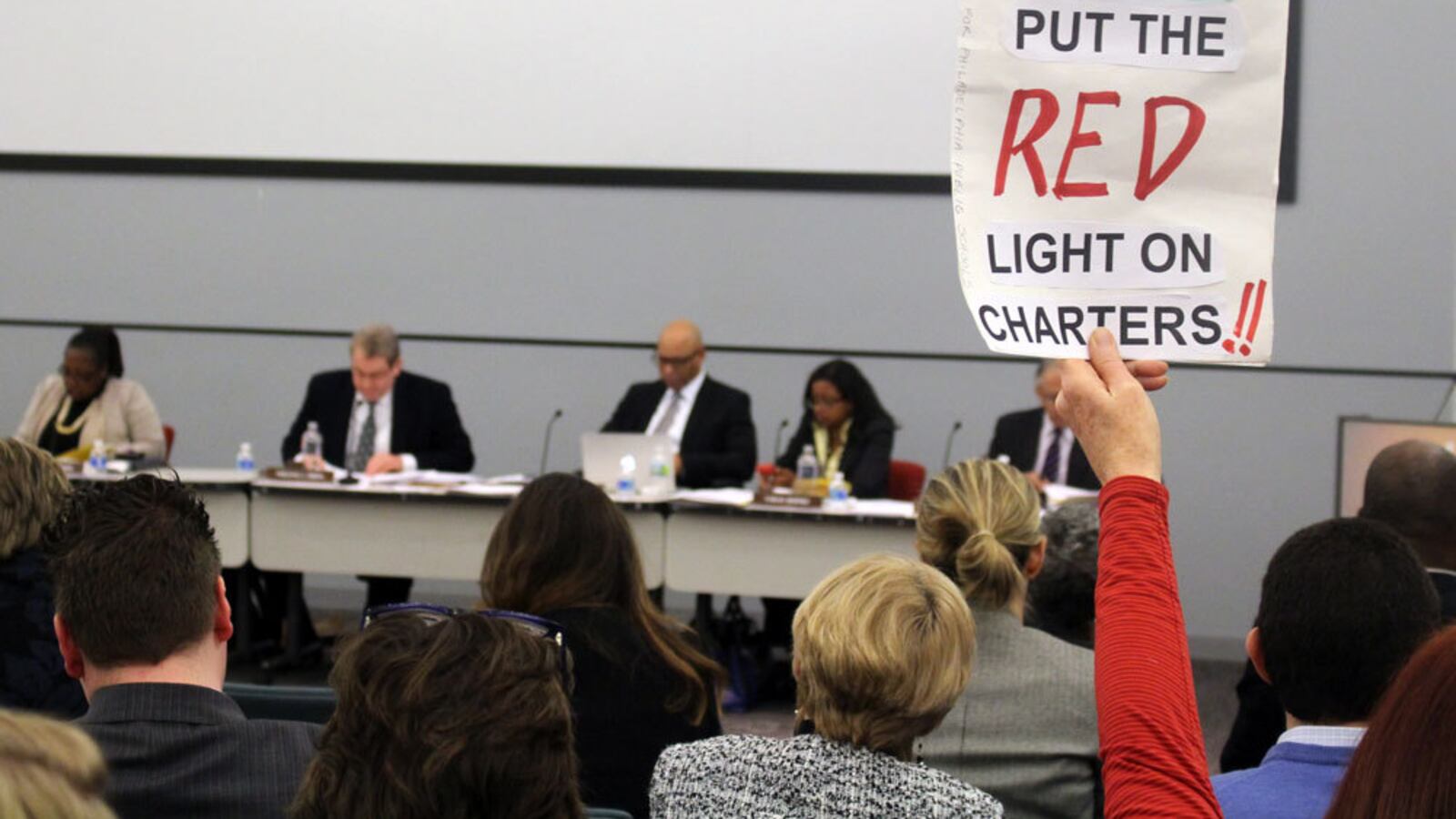 A sign being held up by an audience member at a meeting that reads: Green: put the RED light on charters!