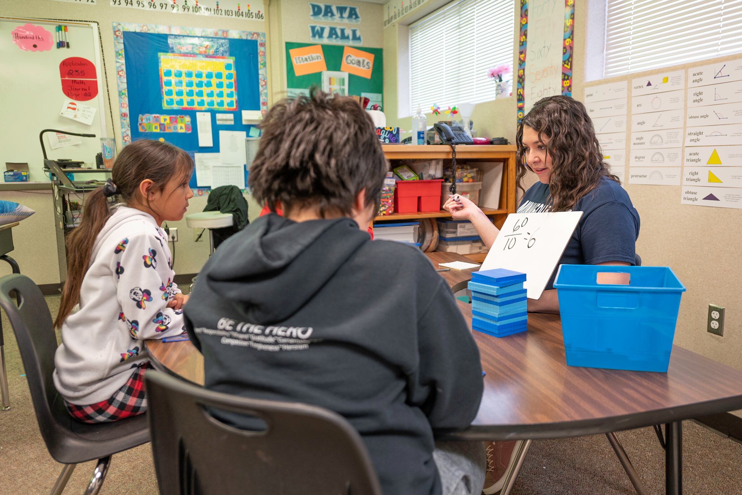A teacher sits a small table holding up a whiteboard so two students can see it.