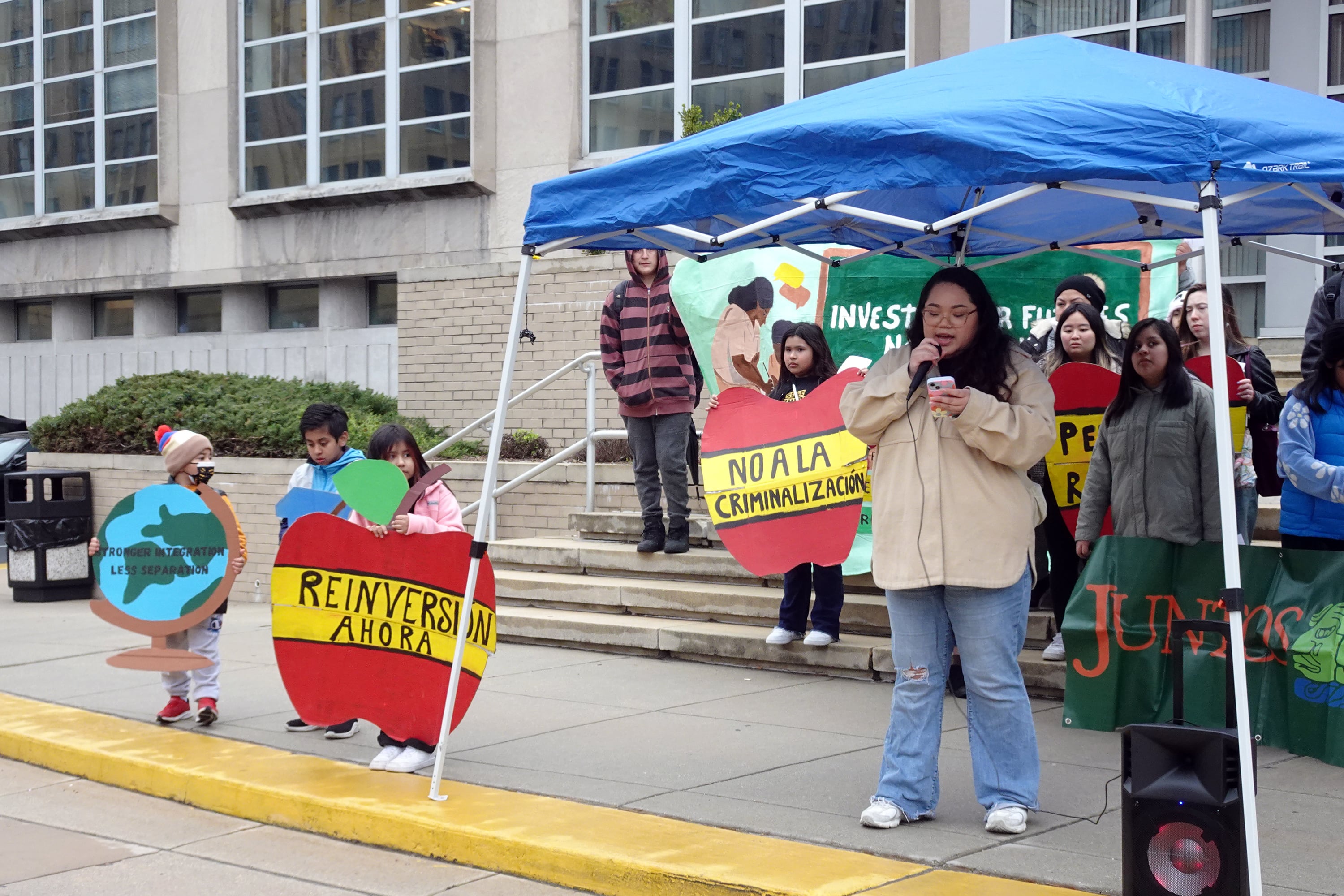A group of young and older people stand out front of a large stone building. Some are holding signs and some are standing under a blue tarp.
