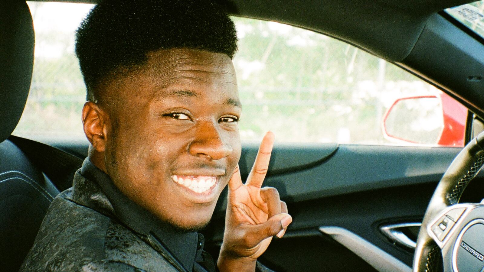 A young man wearing a black tuxedo smiles and flashes a peace sign in the driver’s seat of a car.