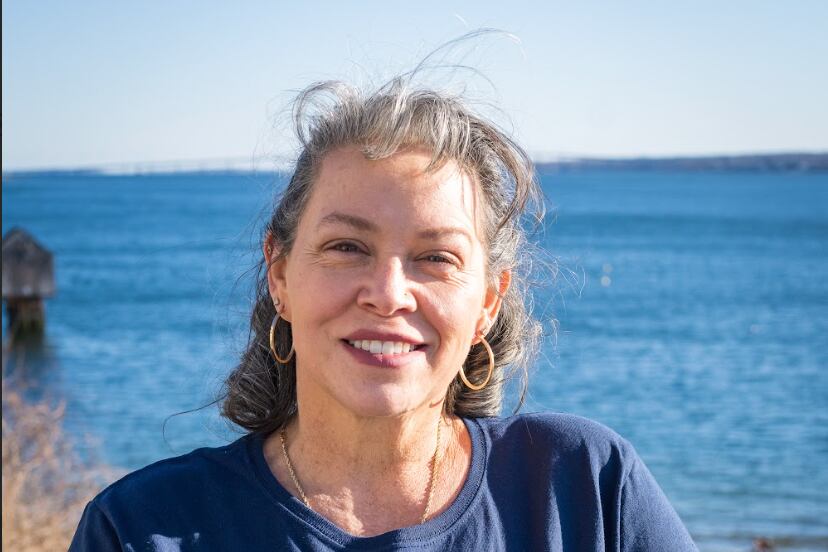 A woman with short hair and wearing a blue shirt poses for a portrait in front of a water view and rocks in the background.