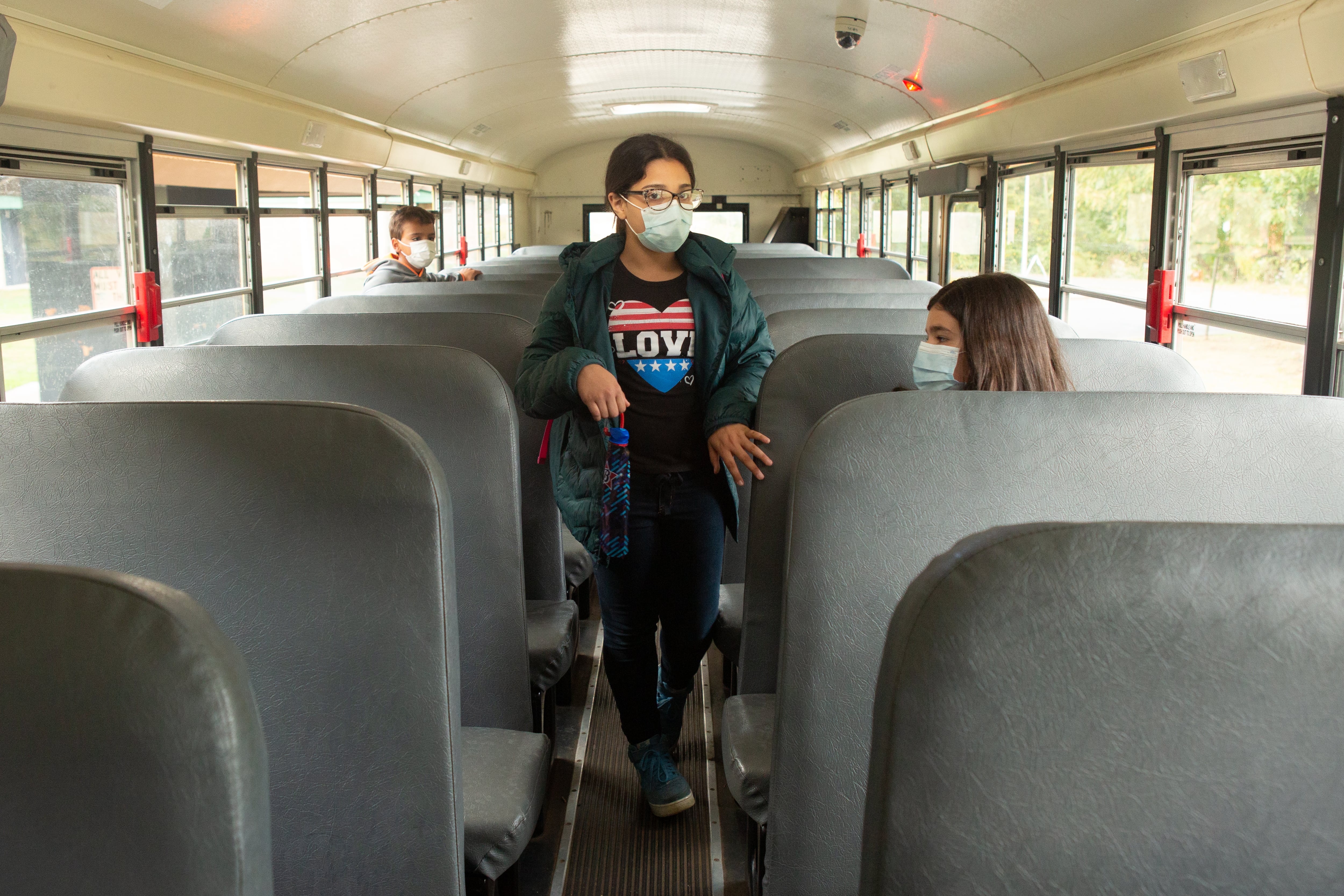 Two fifth graders sit on opposite sides of a school bus while one girl, wearing glasses and a mask and a black T-shirt emblazoned with a red-and-blue heart saying “LOVE” and carrying an umbrella, walks toward the back of the bus.