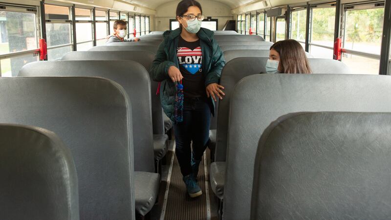 Two fifth graders sit on opposite sides of a school bus while one girl, wearing glasses and a mask and a black T-shirt emblazoned with a red-and-blue heart saying “LOVE” and carrying an umbrella, walks toward the back of the bus.