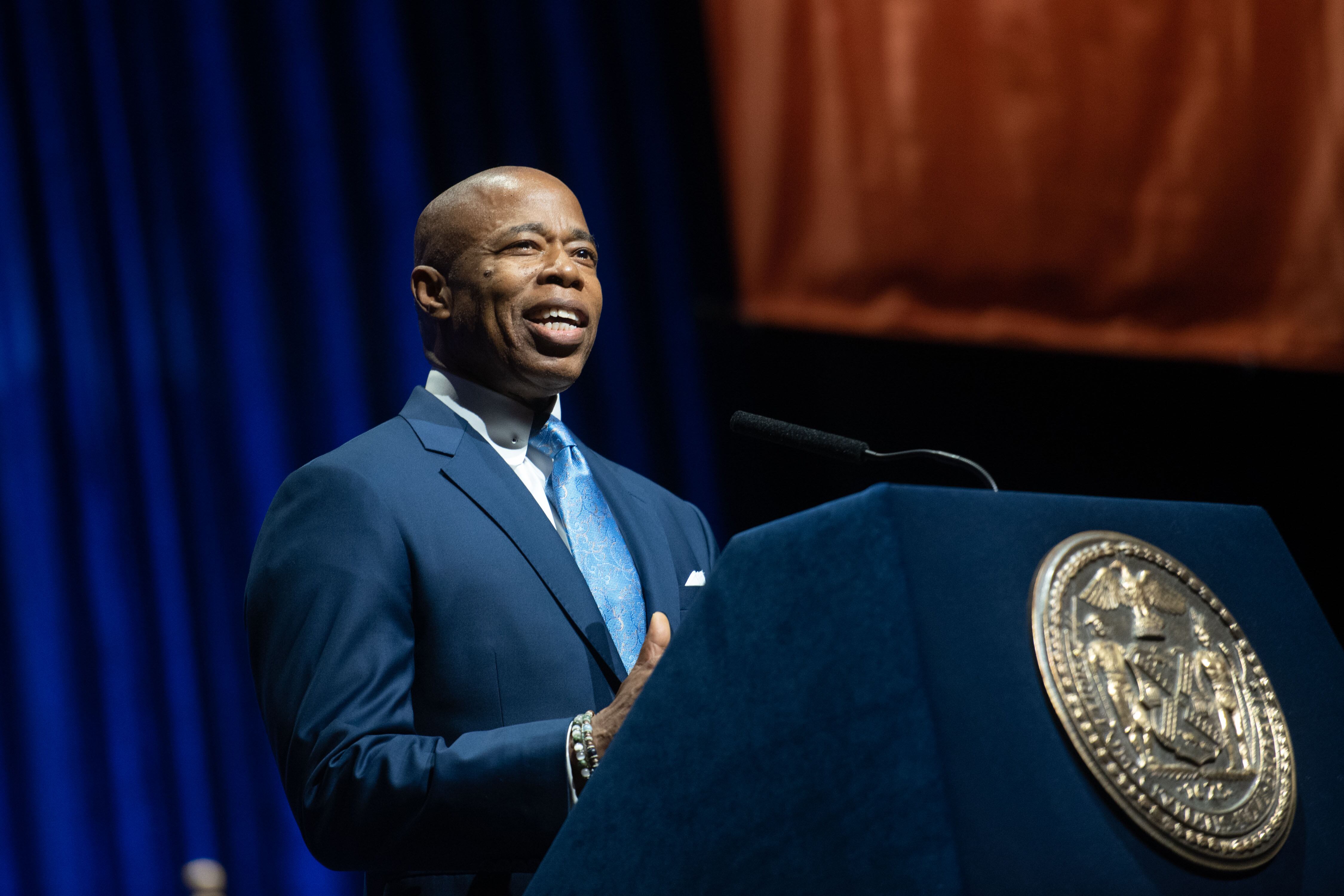 A man wearing a blue suit speaks from a podium with a dark background.