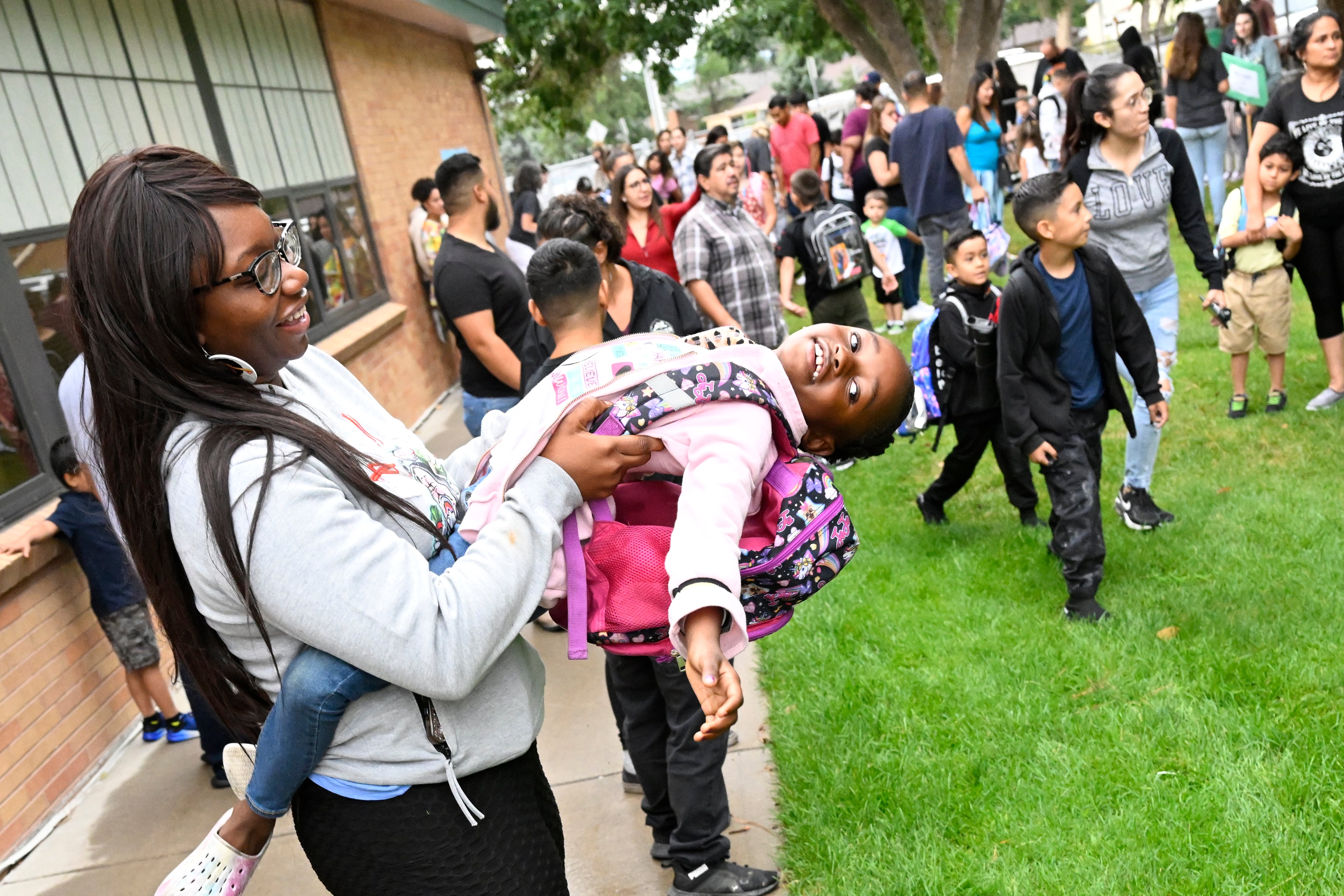 A young girl with her legs wrapped around her mother’s waist leans all the way back with a big smile on her face. Her mother supports her with her arms and smiles down at her lovingly. The girl’s hair is braided with beads and she has a bright pink patterned backpack.