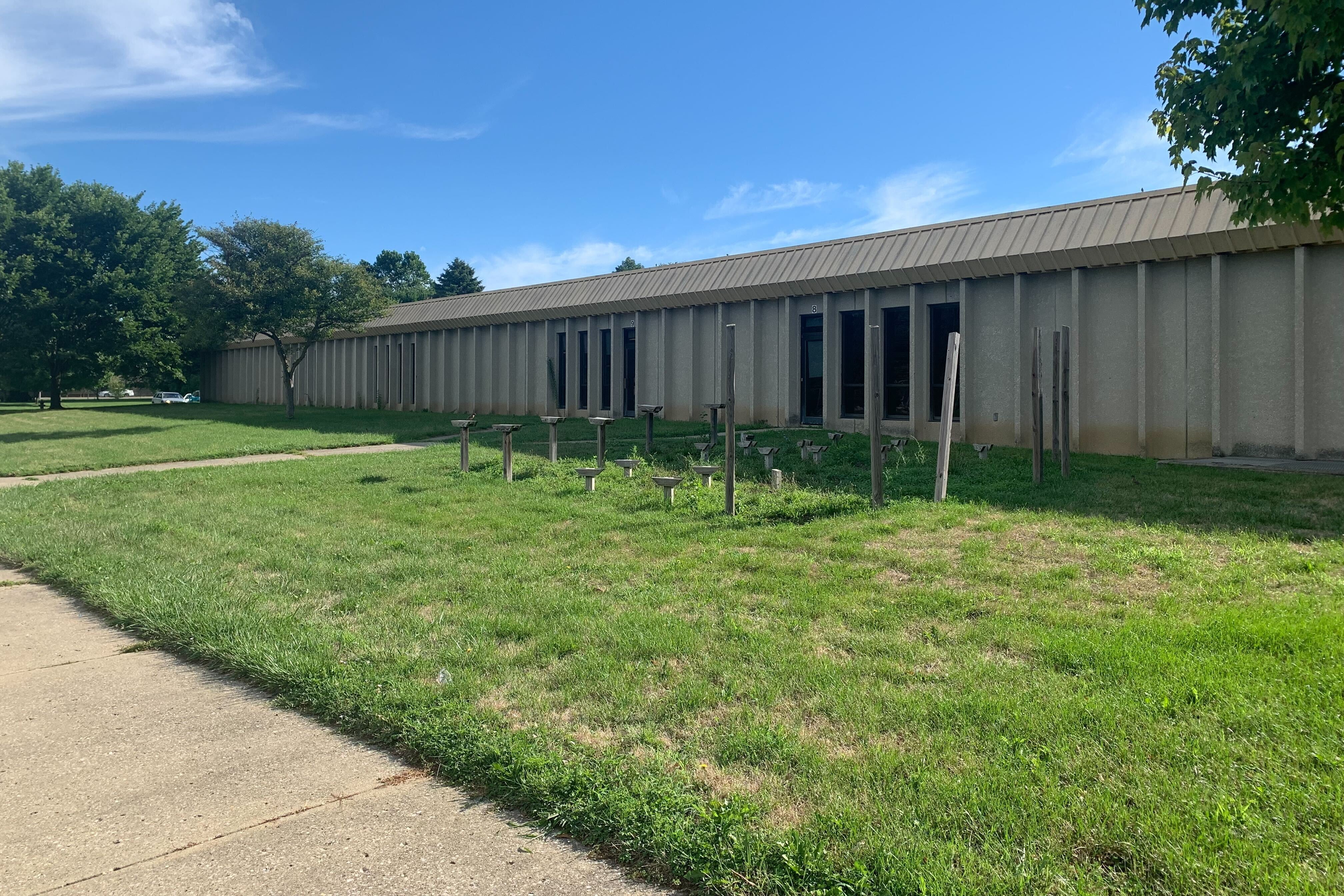 A run down building sits in front of a grass lawn with trees to the building’s right under a blue sky.