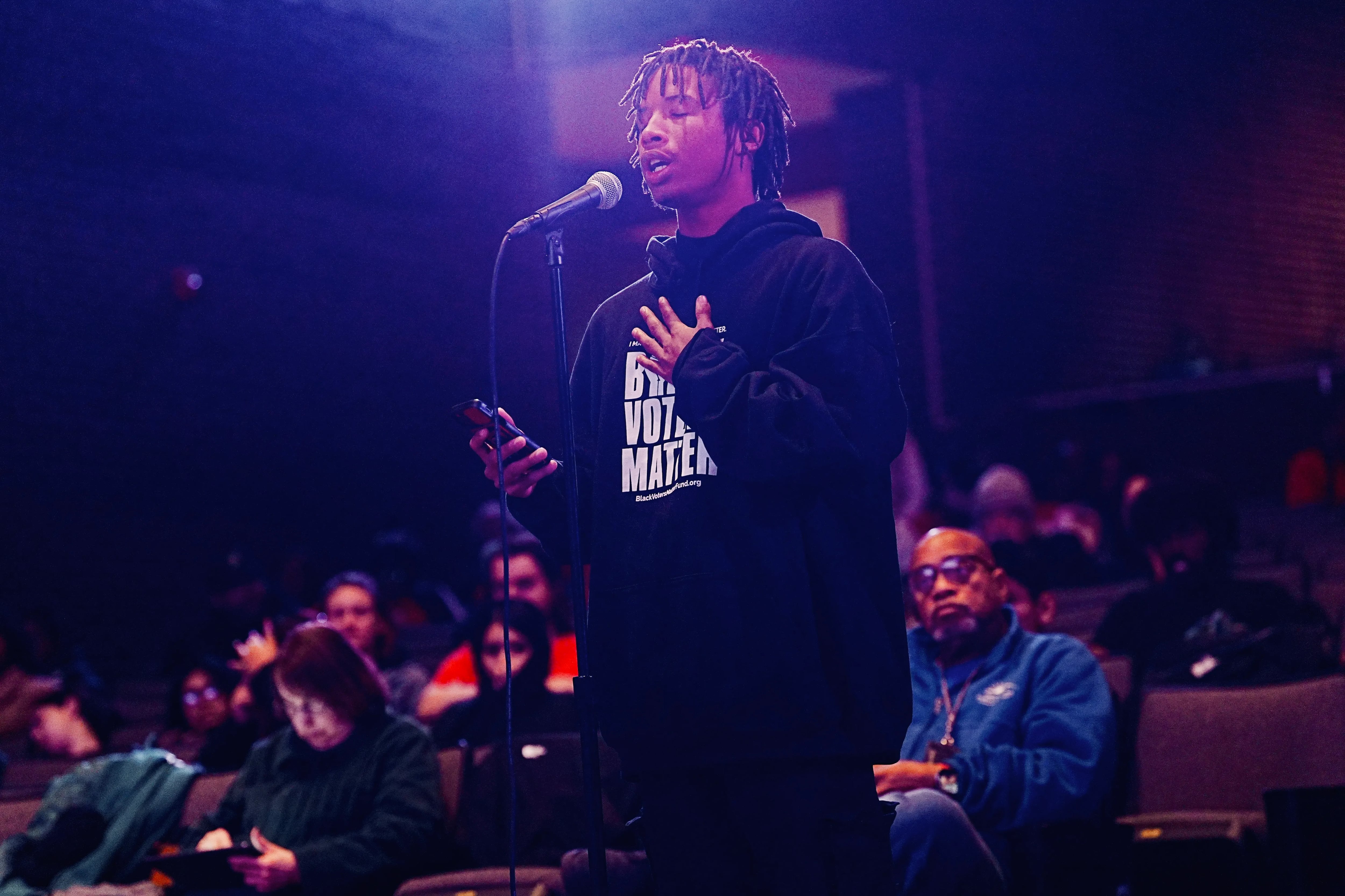 A teenage boy wearing a dark hoodie stands in front of a microphone as he speaks during a meeting.
