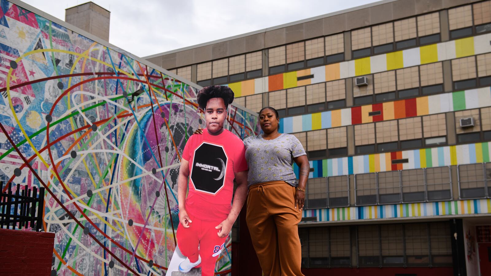A woman in tan pants stands with a cardboard cutout of her son outside of South Philadelphia High School in Philadelphia.