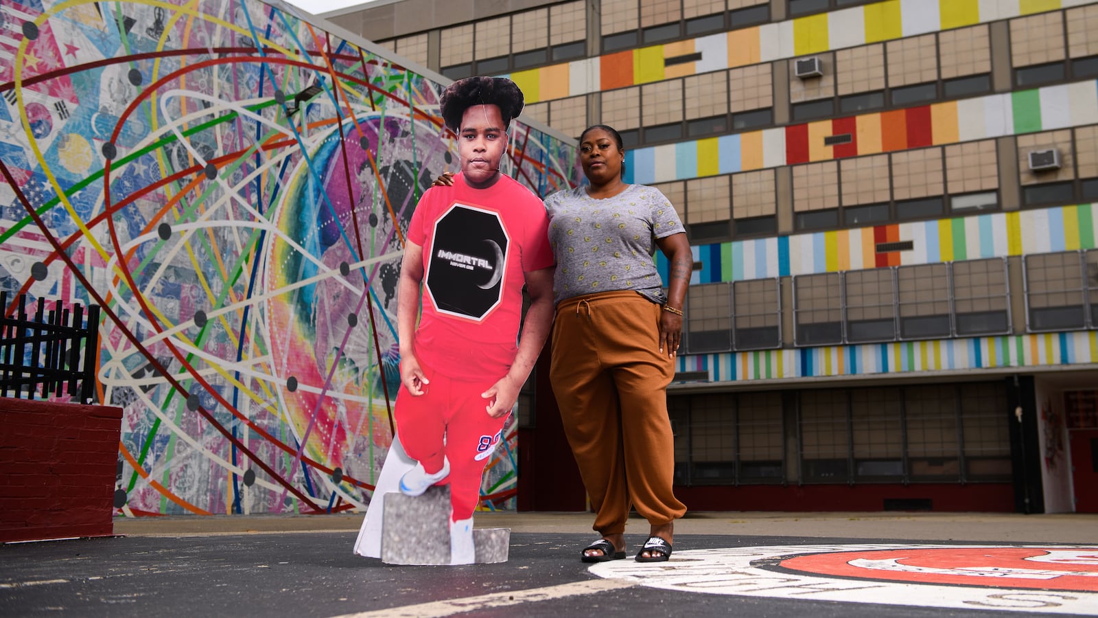 A woman in tan pants stands with a cardboard cutout of her son outside of South Philadelphia High School in Philadelphia.
