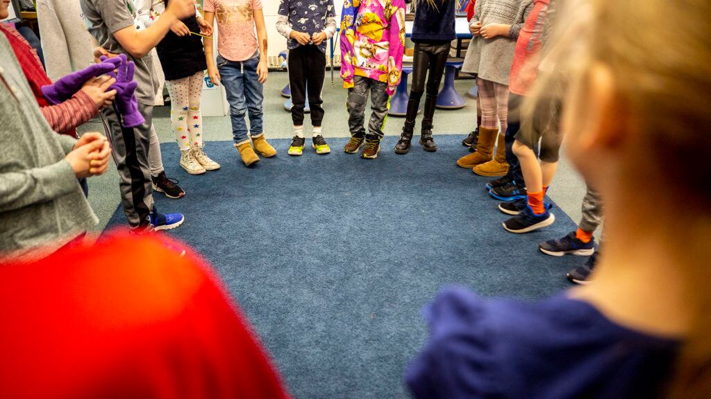 Students standing in a circle on a rug in a room, with their hands clasped, are shown from the waist down