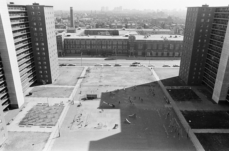 A black and white photograph from 1966 made from above showing a children’s playground between two tall rise buildings on either side and a large three-story, school building in the background.
