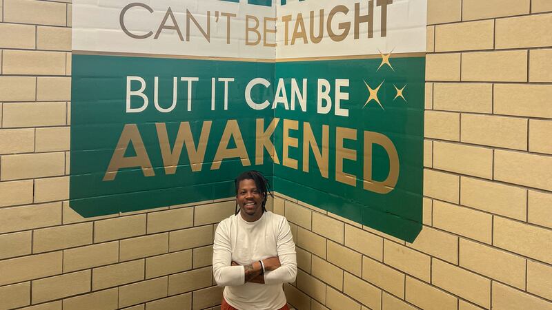 A principal stands under a green and white sign that says “Talent cant’ be taught, but it can be awakened.”