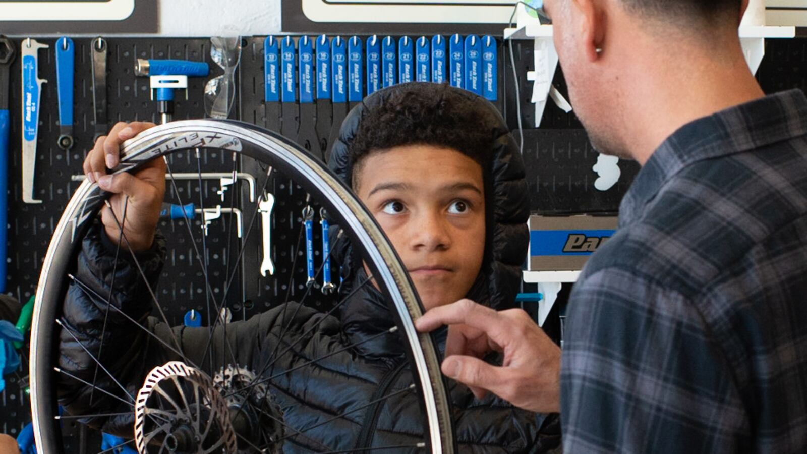 A young teen boy in a black puffy jacket holds a bike wheel aloft while looking at an adult man who is explaining something to him. The man has his back to the camera.